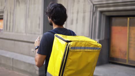 hombre de entrega con mochila amarilla hablar con un cliente en la calle usando auriculares, vista lateral