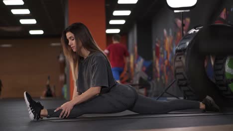 flexible long haired european woman working on extreme leg stretch splits exercise im modern, crossfit gym. determinated girl in sportswear working on her flexibility. side view