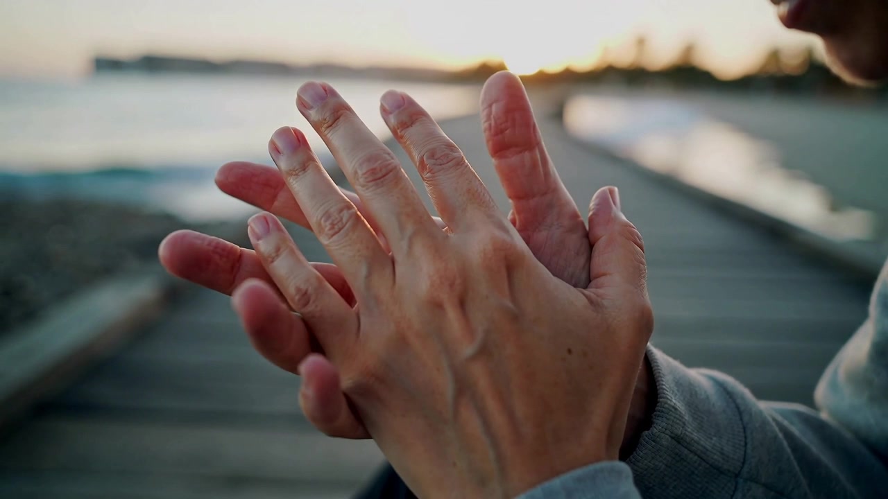 Camera focusing clasped hands, man in hoodie rubbing interlacing fingers on boardwalk by calm sea