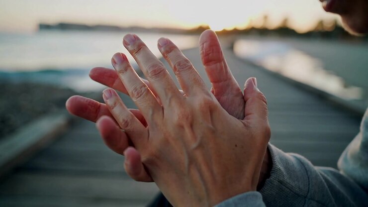 La caméra se concentrant sur des mains jointes, un homme en sweat à capuche frottant ses doigts entrelacés sur une promenade en bois au bord d'une mer calme