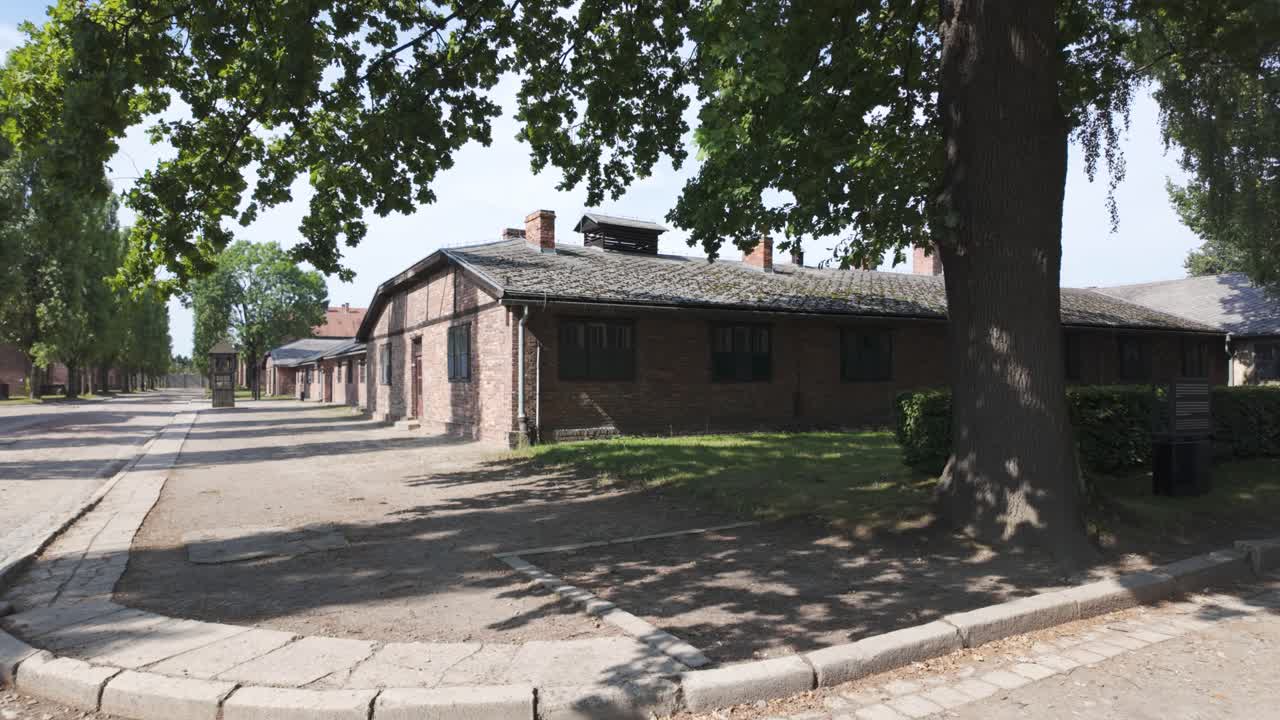 A somber street view of Auschwitz concentration camp in Poland, featuring historic brick buildings and a tree-lined path. Panning Shot