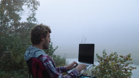 steady shot, a man using a laptop, foggy trees and lake in the background