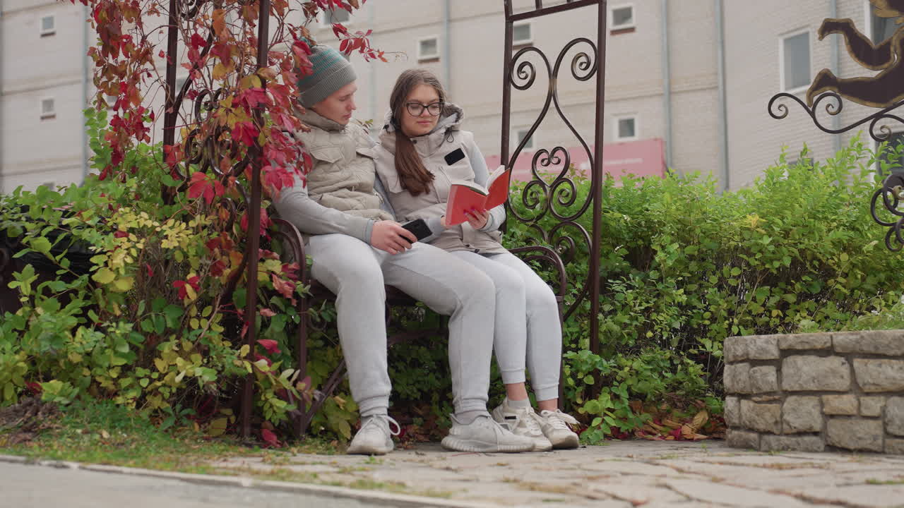 Beautiful moment of lovers holding each other seated outdoor enjoying cool breeze, surrounded by autumn leaves, as woman reads book and man gently embraces her with phone in hand