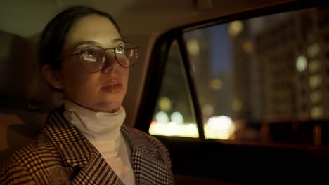 A-confident-brunette-girl-in-round-glasses-and-a-white-and-black-coat-drives-through-the-evening-city-in-the-interior-of-a-modern-car-after-her-successful-business-meeting