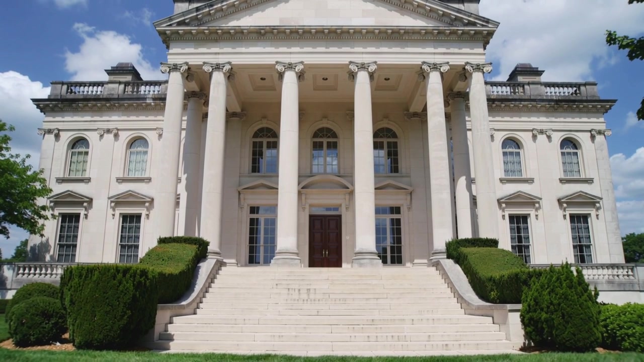 Telecamera in movimento che riprende la facciata di una villa neoclassica su un prato, mostrando colonne, gradini e porte.