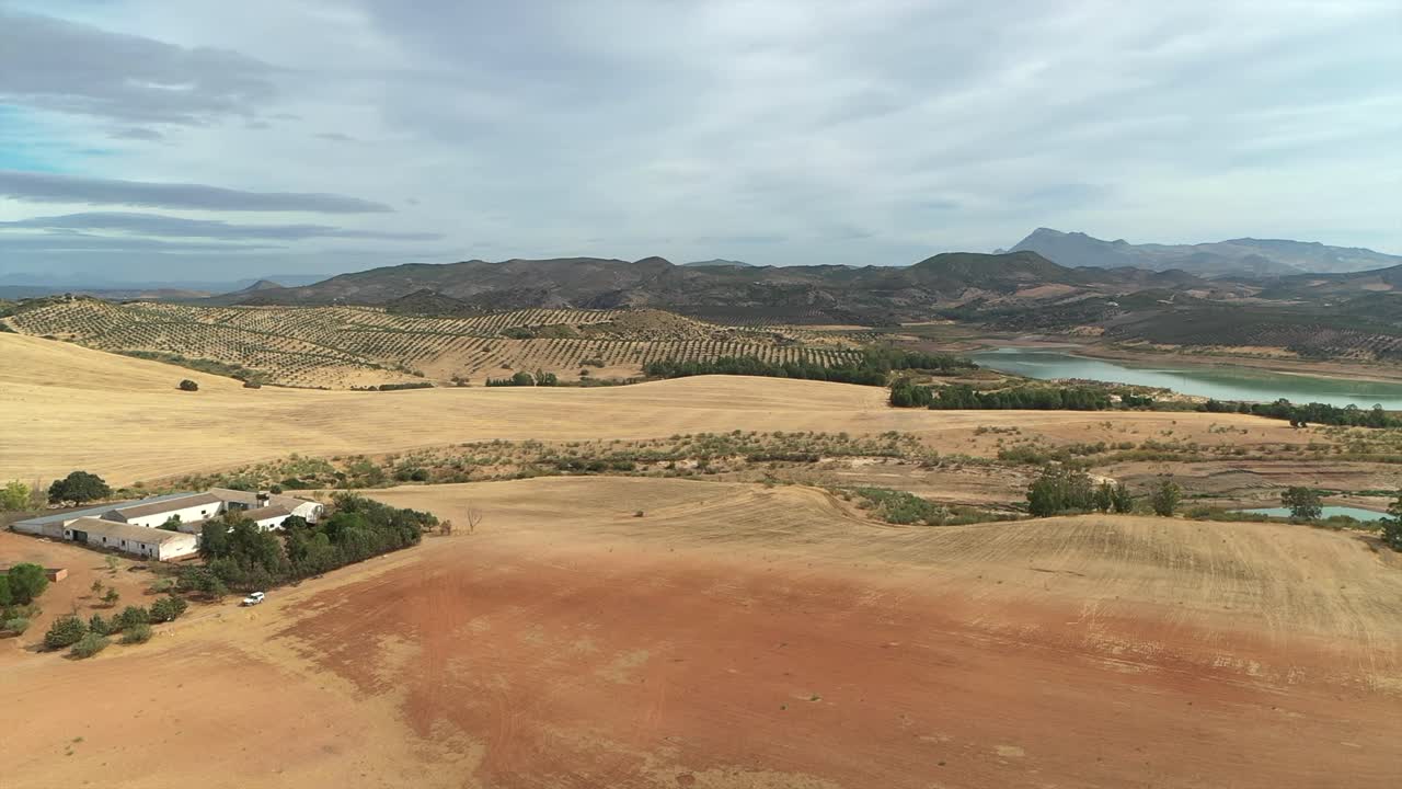 Farmhouse surrounded by vast, dry agricultural fields and rolling hills near a reservoir in rural Andalusia, Spain, aerial