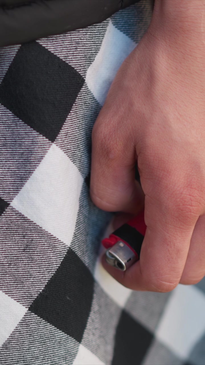 Primer plano de una mano sosteniendo un encendedor rojo contra una tela a cuadros con movimiento mínimo. El enfoque permanece en el detalle de la textura y la interacción entre la piel, el objeto y el material. Iluminación suave.
