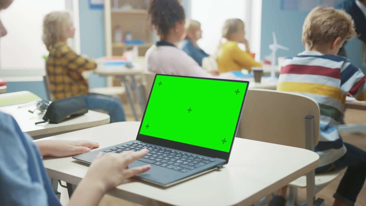 Elementary School Classroom: Over the Shoulder View of a Child Using Green Mock-up Screen Laptop in Class. In the Background Teacher Explains Lesson to Diverse Class