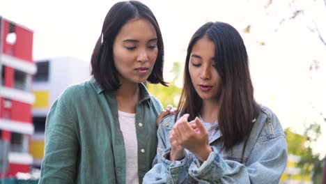 two young japanese girls looking their nails and talking about manicure while standing outdoors in the street