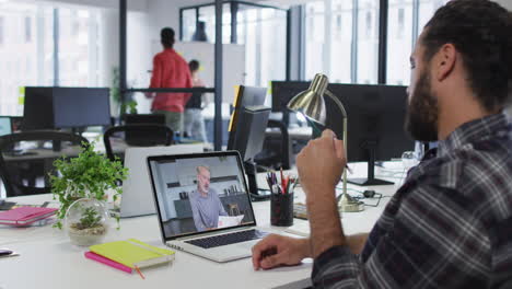 hombre de negocios biracial sentado en un escritorio usando una computadora portátil haciendo una llamada de video con un colega masculino