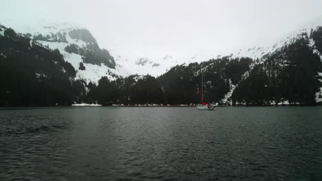 Drone-approaching-a-sailboat-anchored-in-middle-of-snowy-islands-of-cloudy-Alaska