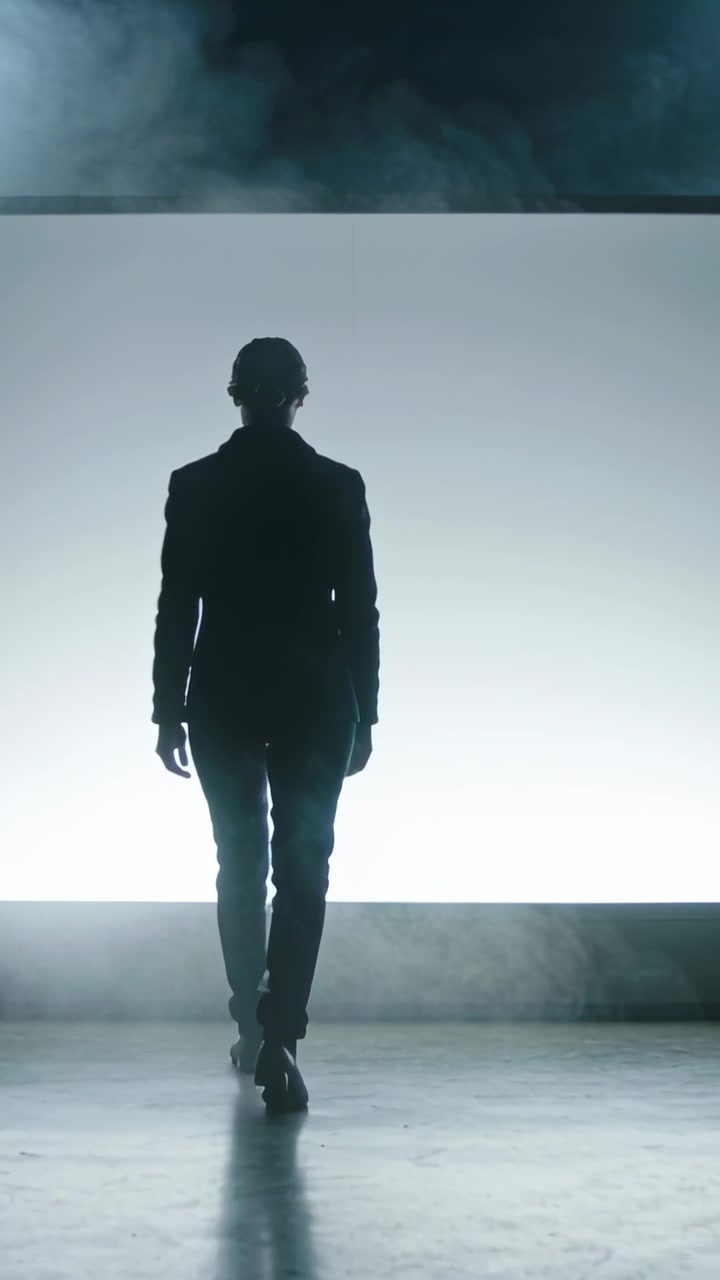 Video verticale: Modello che entra in scena in giacca, camminando verso un pannello retroilluminato sul palco, con nebbia che sale.