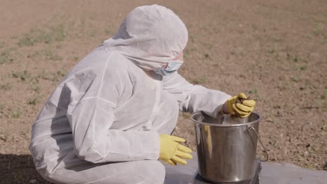 Person-in-protective-suit-in-field-soil-sampling,-bucket-with-ground-sample