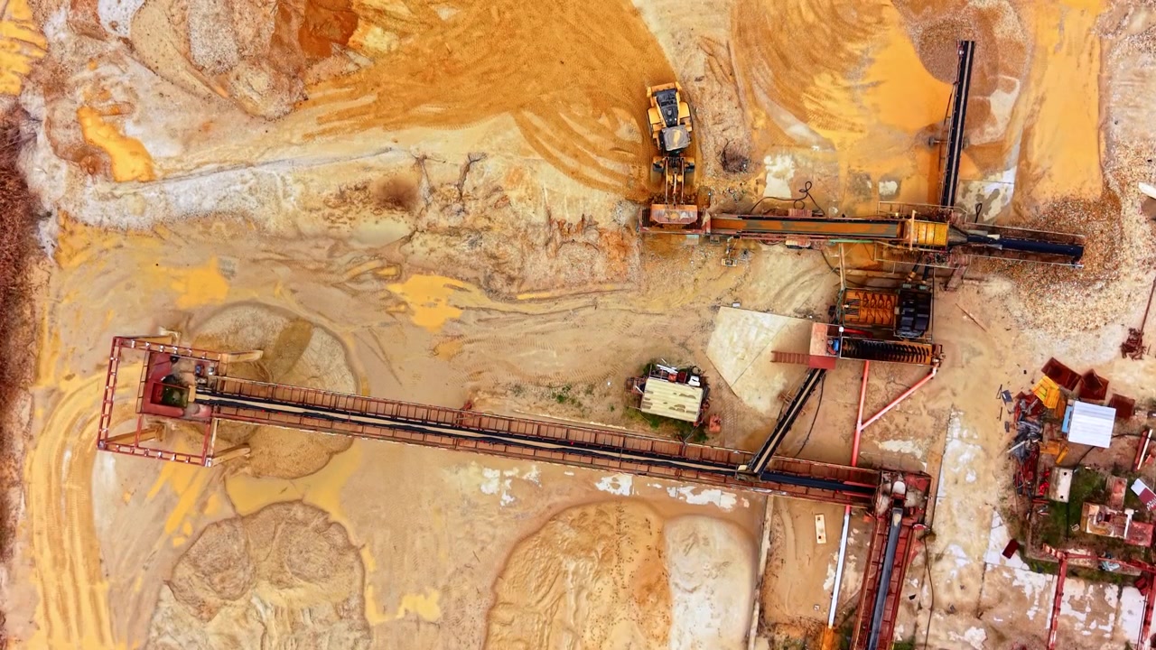 Conveyor belt and loader at sand processing site. Overhead view captures a wheel loader beside a long red conveyor and machinery frame.