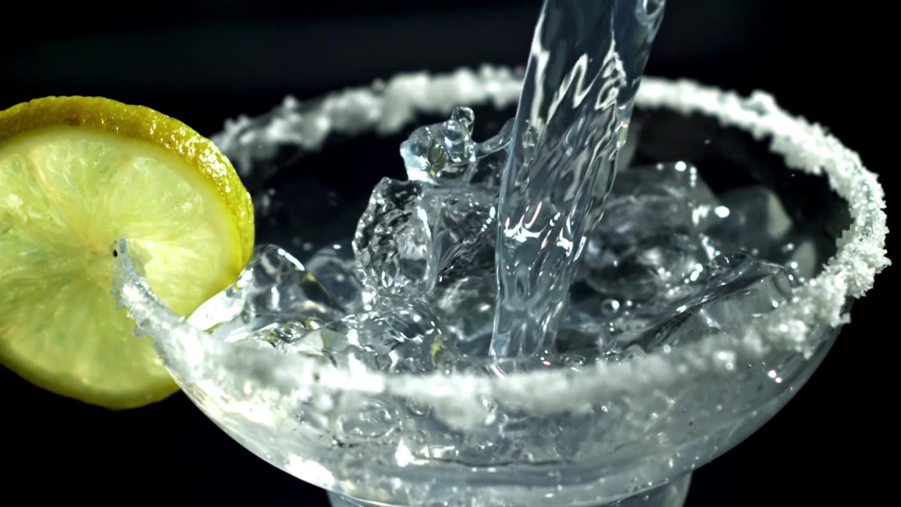 Liquido trasparente che si versa in un bicchiere da cocktail con bordo salato, ghiaccio e limone al rallentatore