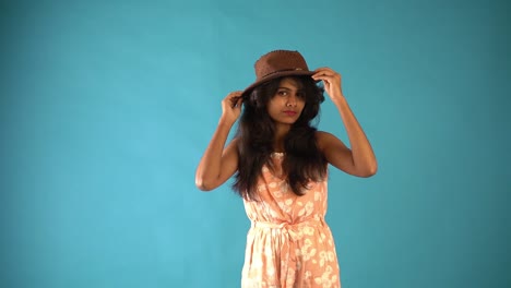 Ein-Junges-Indisches-Mädchen-In-Orangefarbenem-Kleid-Mit-Braunem-Hut,-Das-Auf-Einem-Isolierten-Blauen-Hintergrund-Steht