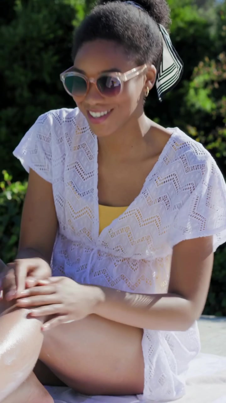 Vertical video: Hand rubbing woman applying sunscreen on legs at patio, crochet cover-up for sun