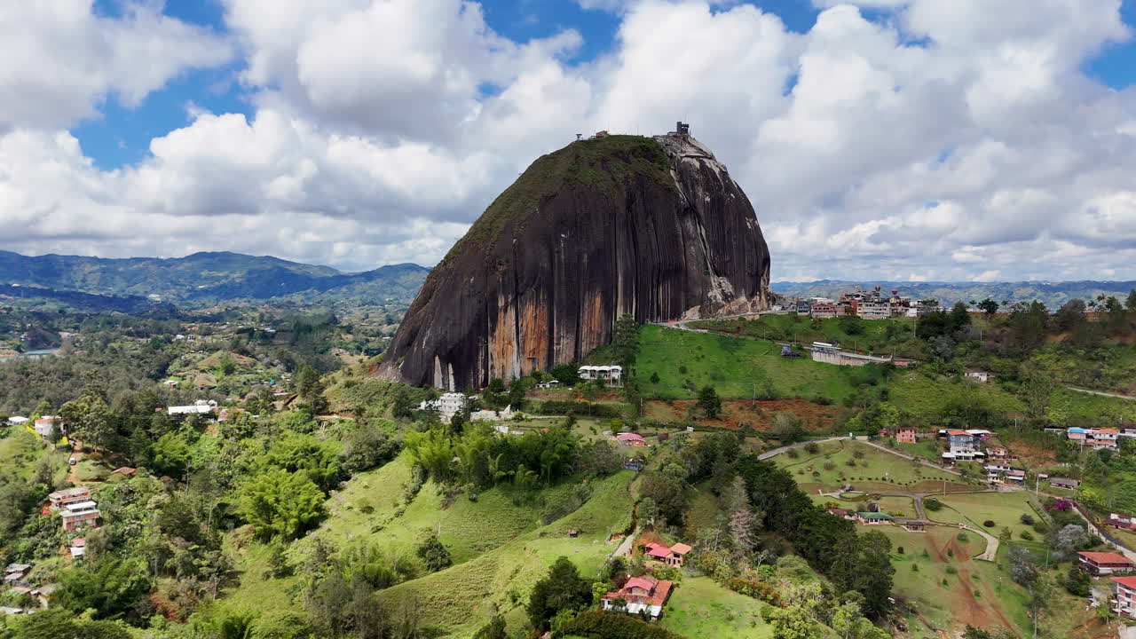 Perspectiva de drone a média distância da icônica Rocha El Peñol (La Piedra del Peñol). Tomada cinematográfica mostrando a escala do maciço monólito granítico elevando-se sobre a paisagem de Antioquia.