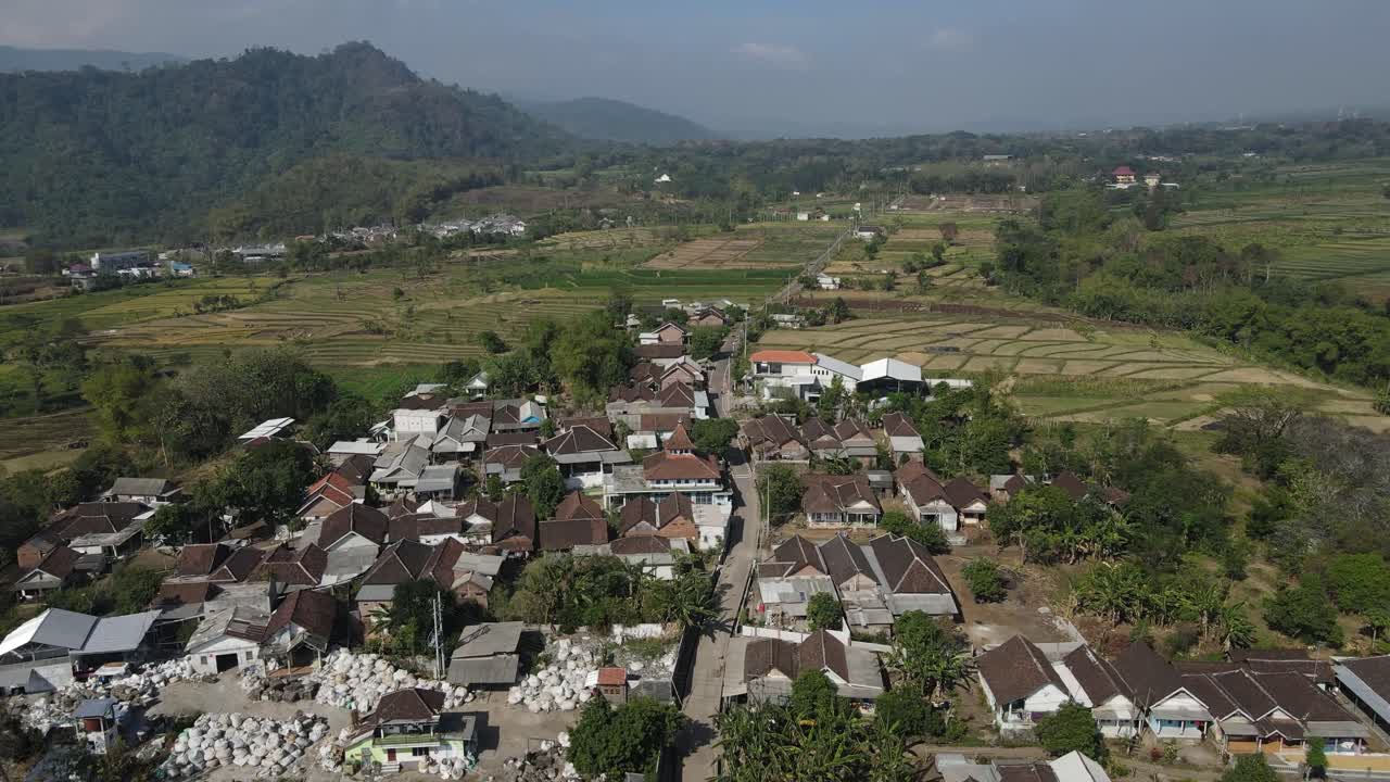 Scenic aerial shot of rural Indonesian town featuring a natural pond, clustered rooftops, farmland, and tropical greenery with mountain landscape in the background