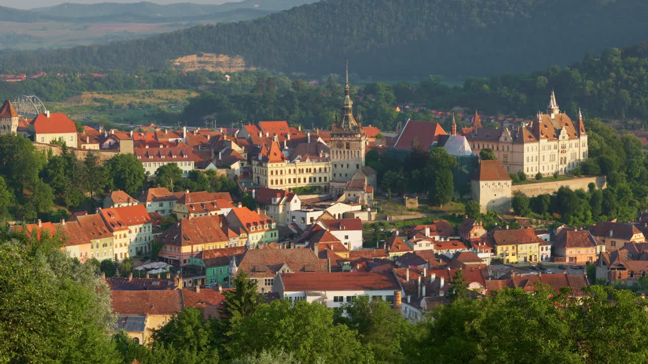 Ujęcie panoramiczne porannej Sighișoary w Rumunii. Poranne słońce oświetla pomarańczowe dachy starego domu i ratusza, nad którymi latają stada gołębi.