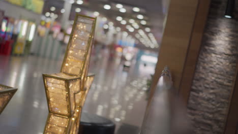 primer plano de intrincados marcos dorados con patrones detallados que brillan cálidamente, rodeados de luces blancas parpadeantes, el fondo presenta un interior borroso del centro comercial con pisos reflectantes y efectos bokeh suaves