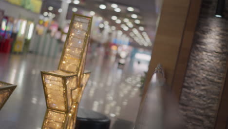 close-up of intricate gold frames with detailed patterns glowing warmly, surrounded by blinking white lights, background features a blurred mall interior with reflective floors and soft bokeh effects