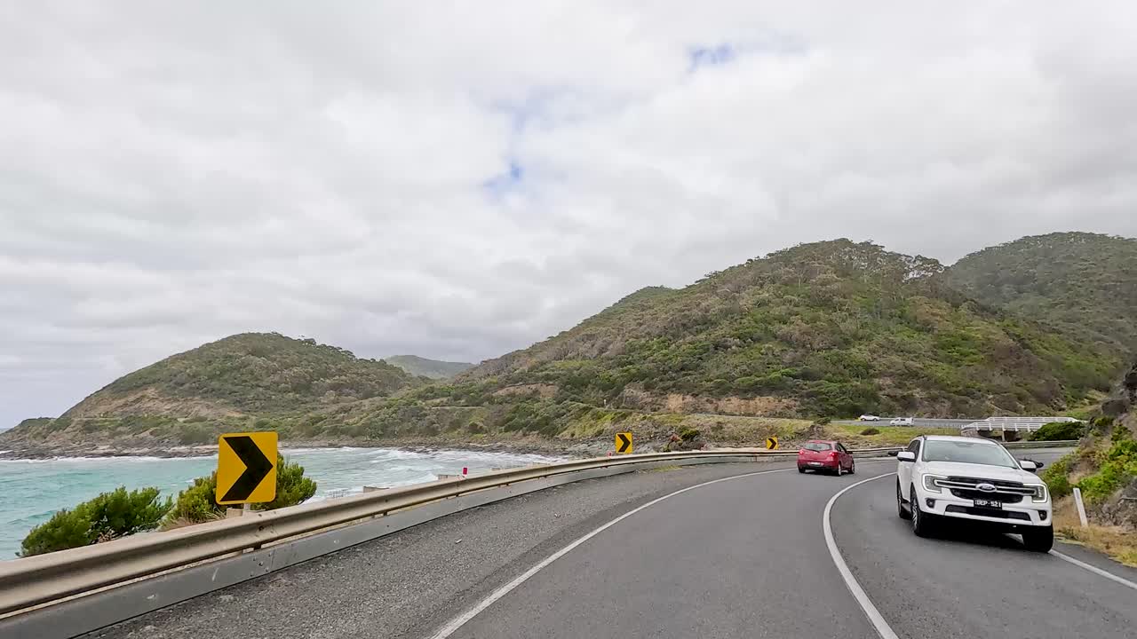Cars travel a coastal road with ocean views and lush hills under cloudy skies, capturing the essence of a scenic journey
