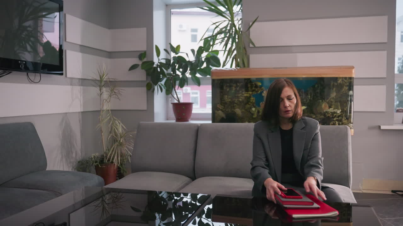 Young professional sitting on gray couch, preparing documents, holding red notebook, focused on organizing work in modern office with aquarium and plants in background