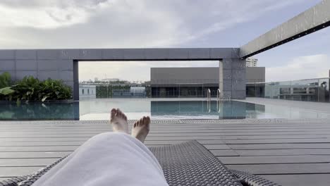 someone resting on the poolside outdoor lounger overlooking at calm rooftop pool on a beautiful sunny day, point of view holiday vacation shot