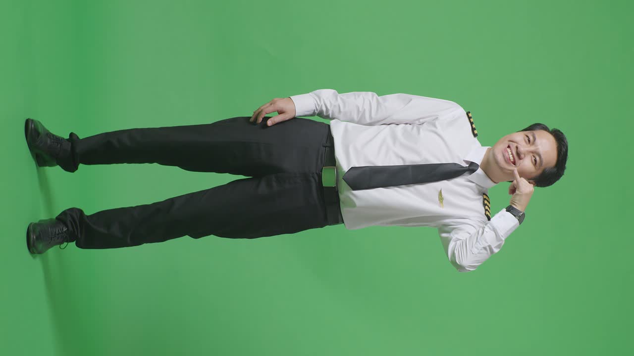아시아 남성 조종사가 미소 짓고 나를 불러서 녹색 스크린 배경 스튜디오에 서있는 동안 카메라에 제스처를합니다.