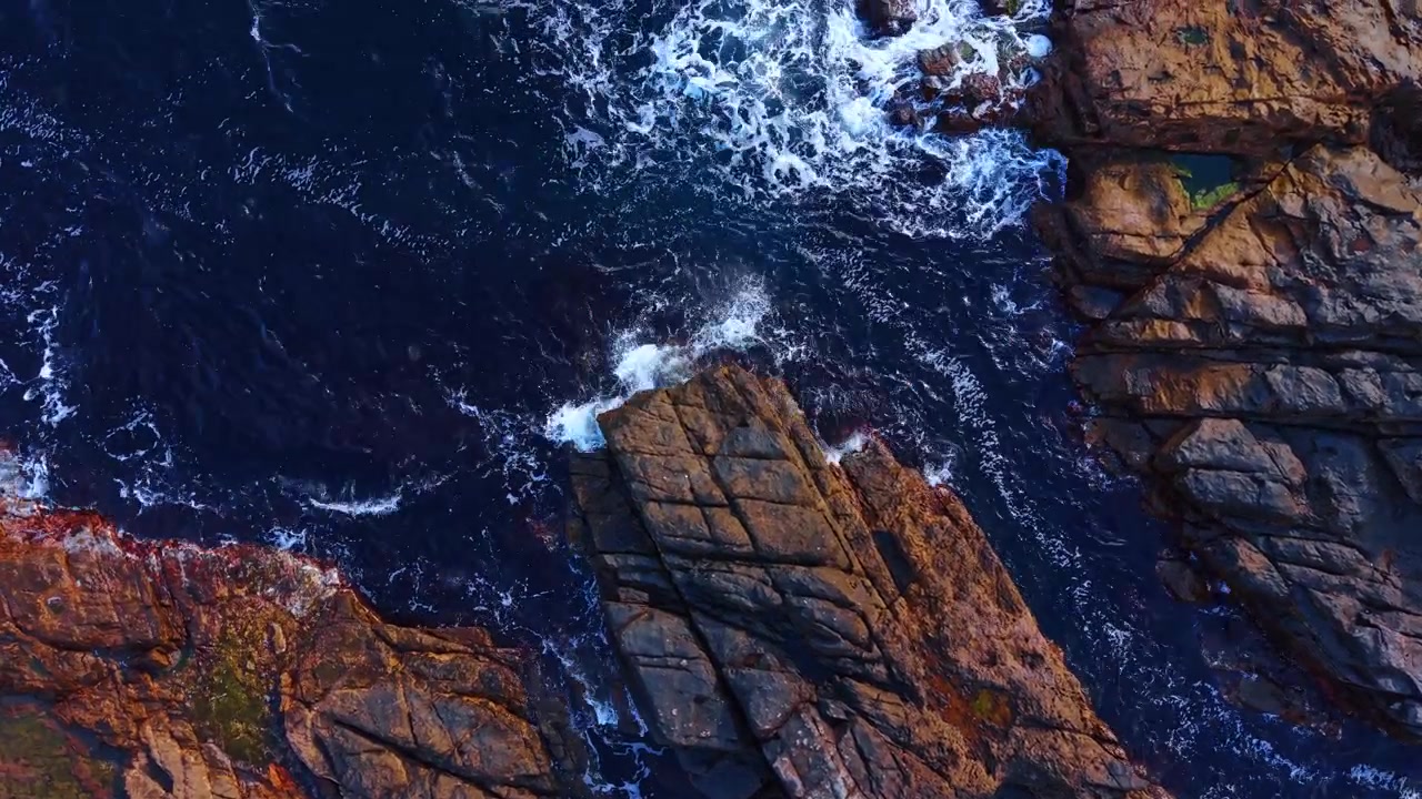 Diagonale Felsplatten, von dunklen Meereswellen von oben umspült. Zerbrochenes Sedimentgestein mit weißer Brandung und rostfarbenen Kanten in tiefblauem Wasser.