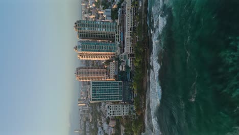 Toma-Aérea-Vertical-Del-Mar-Caribe-Ondulado-Y-Avenida-George-Washington-En-Santo-Domingo-Con-Horizonte-Durante-La-Puesta-De-Sol-Dorada