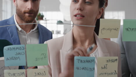 corporate-business-people-using-sticky-notes-brainstorming-problem-solving-strategy-on-glass-whiteboard-team-leader-woman-showing-solution-for-project-deadline-in-office-meeting