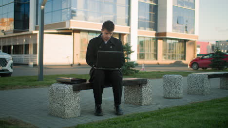 businessman seated on outdoor bench operating laptop near modern office building surrounded by greenery and urban infrastructure, red car and architectural reflections in background