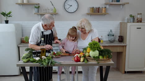 Elderly-grandparents-in-kitchen-teaching-grandchild-girl-how-to-cook-salad,-chopping-red-pepper