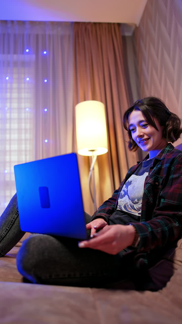 Approaching a relaxed Caucasian lady sitting on a large sofa. Girl looks at her laptop smiling. Low angle view. Christmas tree and lights at backdrop. Vertical video.
