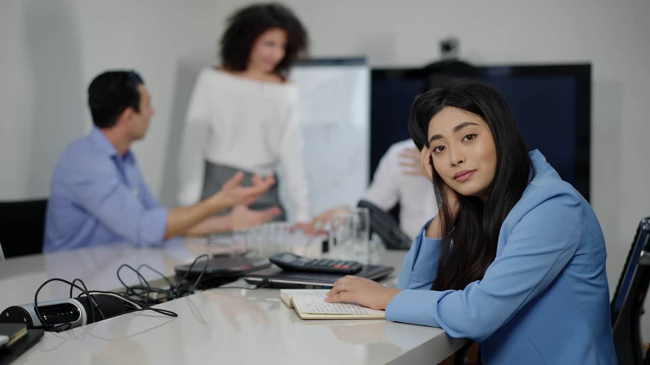 Charming slim Asian businesswoman sitting at conference table shaking head as blurred colleagues arguing at background. Stressed young woman posing in office during brainstorming. Teamwork concept.