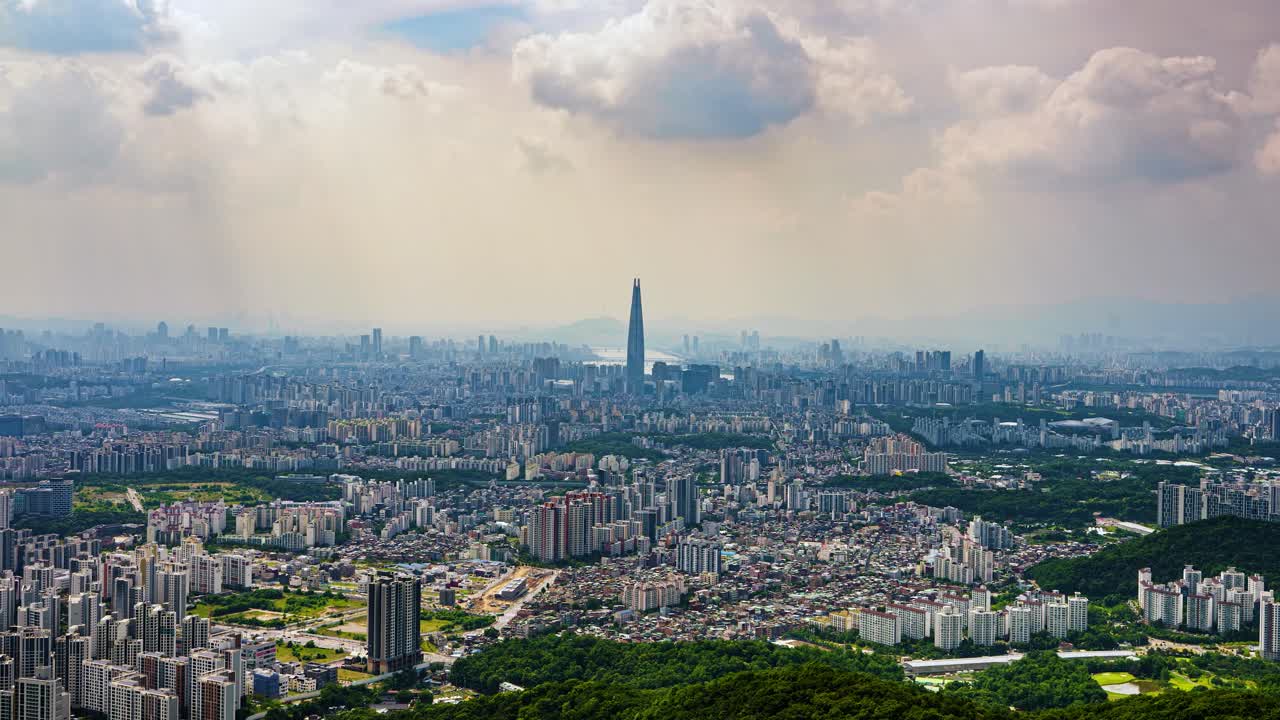 A dramatic timelapse shows clouds casting moving shadows over the vast urban sprawl of Seoul, South Korea, with the iconic Lotte World Tower standing tall in the center of the cityscape.