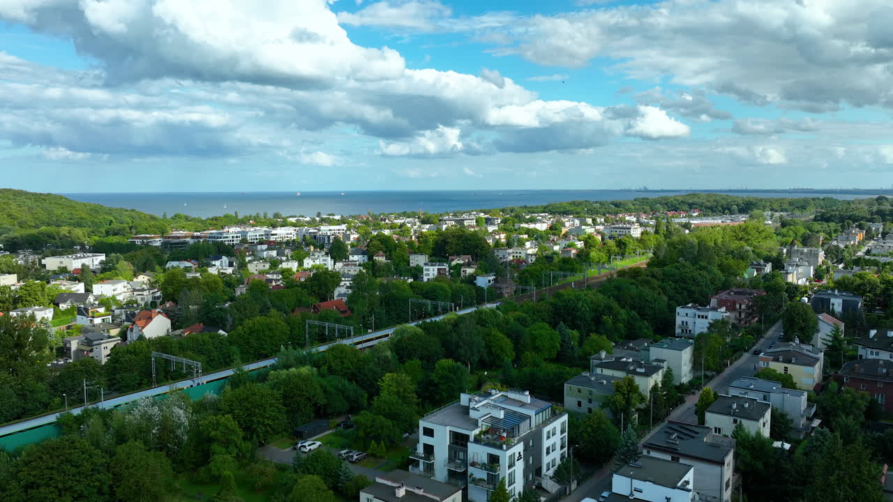 Gdynia - Aerial view of a residential neighborhood with low-rise buildings surrounded by lush green trees. In the distance, the sea meets the horizon under a partly cloudy sky