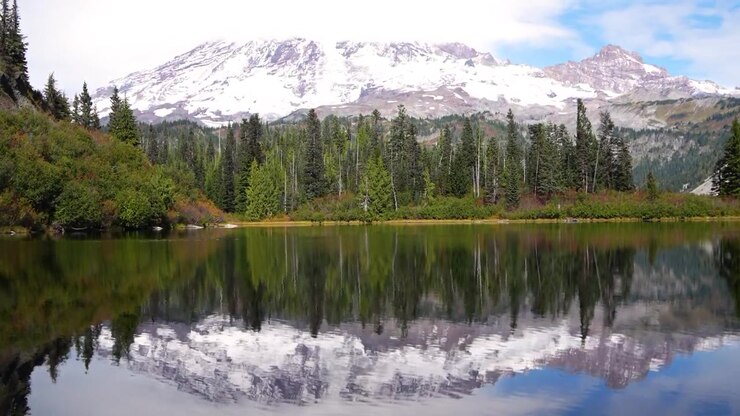 Il Monte Shuksan che si erge sopra una foresta sempreverde e si riflette sulla superficie calma del Lago Picture. Pendii coperti di neve e cieli parzialmente nuvolosi creano un suggestivo scenario alpino