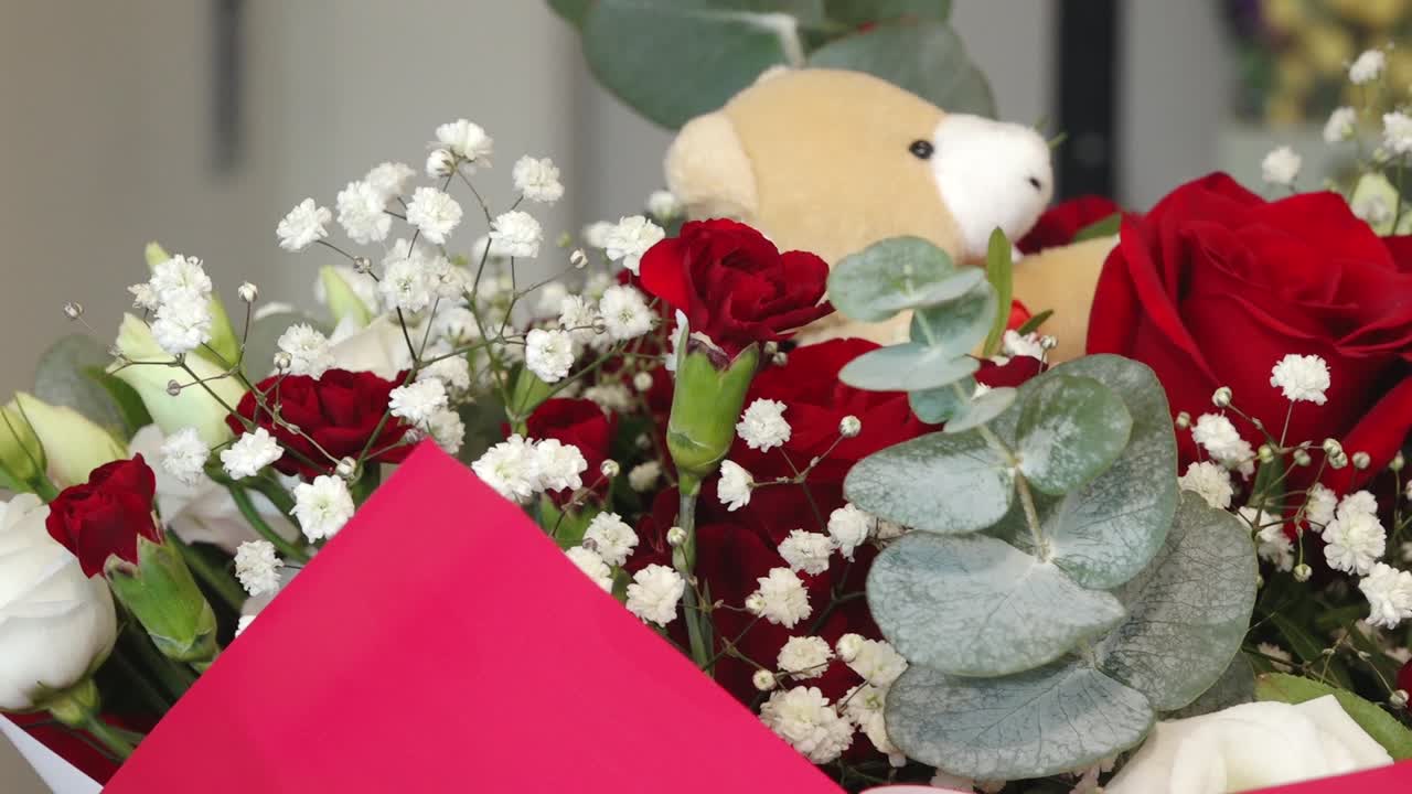 a soft teddy bear nestled among vibrant red roses, white baby's breath, and green foliage. Romantic floral arrangement with a warm, affectionate mood