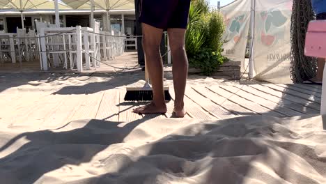 Young-person-sweeping-sand-off-the-wooden-boardwalk-at-a-beach-bar