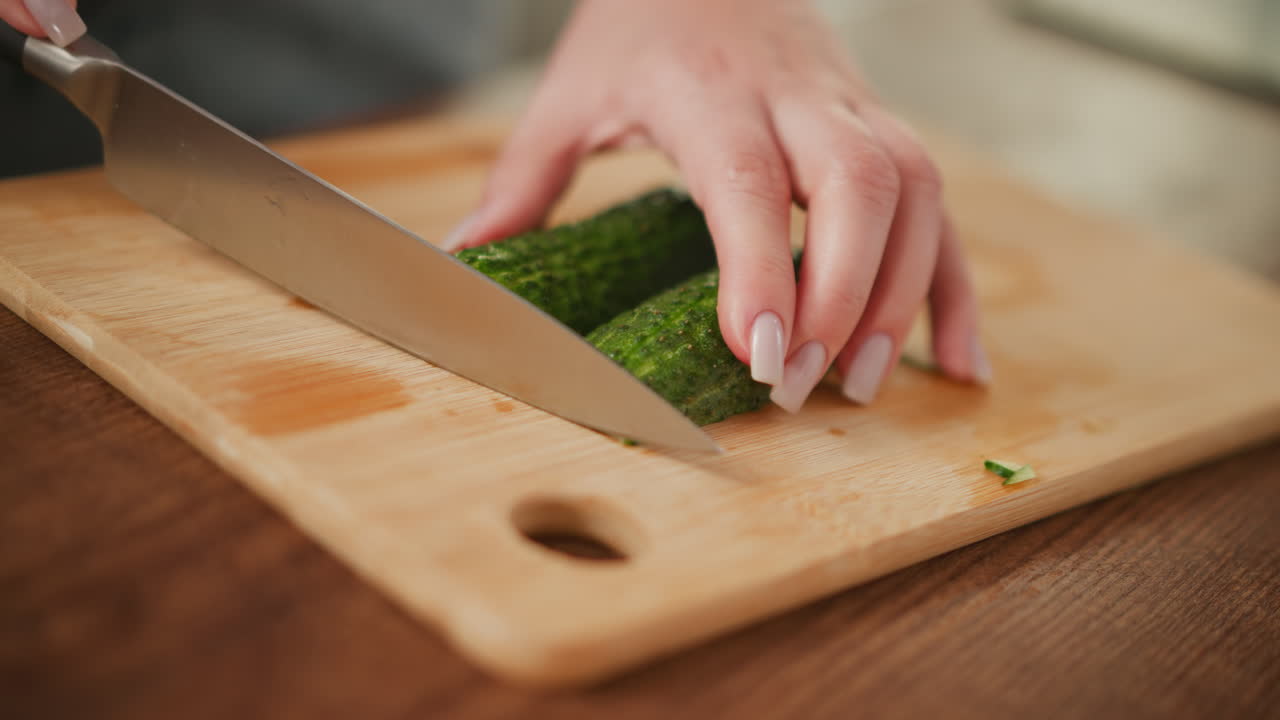 Close up of person holding fresh cucumber and carefully slicing it into two equal parts on wooden cutting board, with focus on hands, knife, and clean kitchen prep environment
