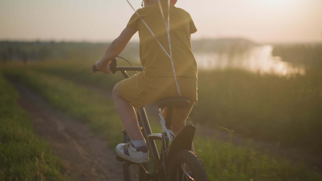 A young boy, dressed in yellow, pedals his bicycle through a grassy field at sunset. The scene captures a vibrant red balloon tied to the back of his bike, with the warm sunlight