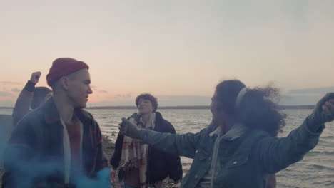 group of teenage friends dancing while holding colored flare lights on the seashore