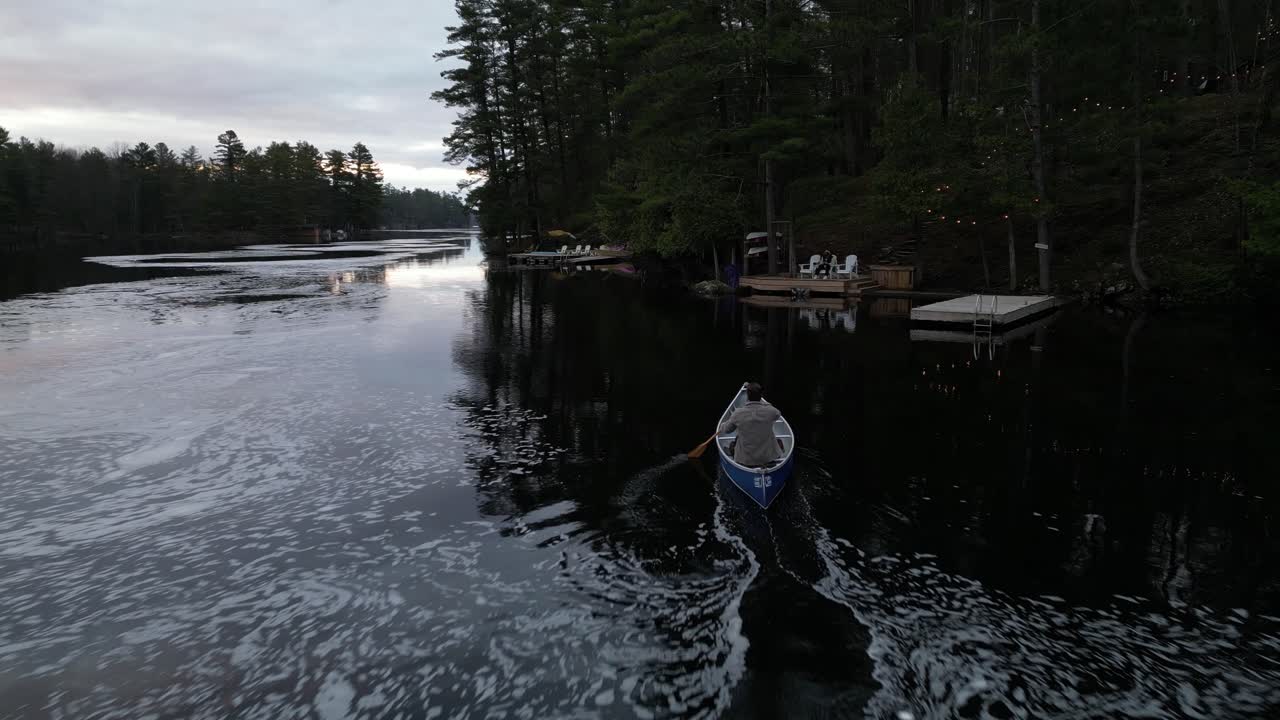 Person rowing a canoe on a calm river by forested banks at dusk, with reflections of trees and dock lights on the water