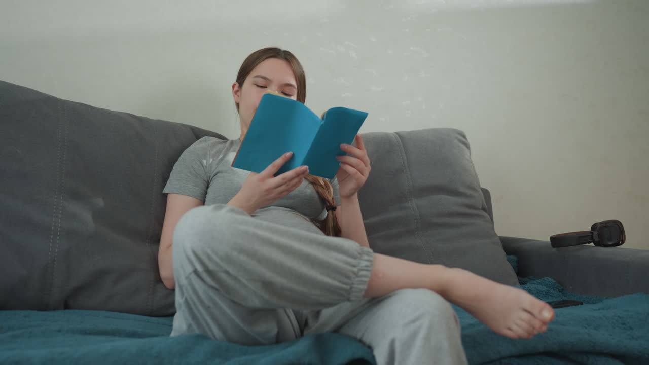 Futura mamma che legge sul divano, gambe incrociate, libro blu sulla genitorialità tra le mani, espressione concentrata, abbigliamento casual da casa, luce soffusa, che rivede liste di controllo e consigli, preparandosi all'arrivo.