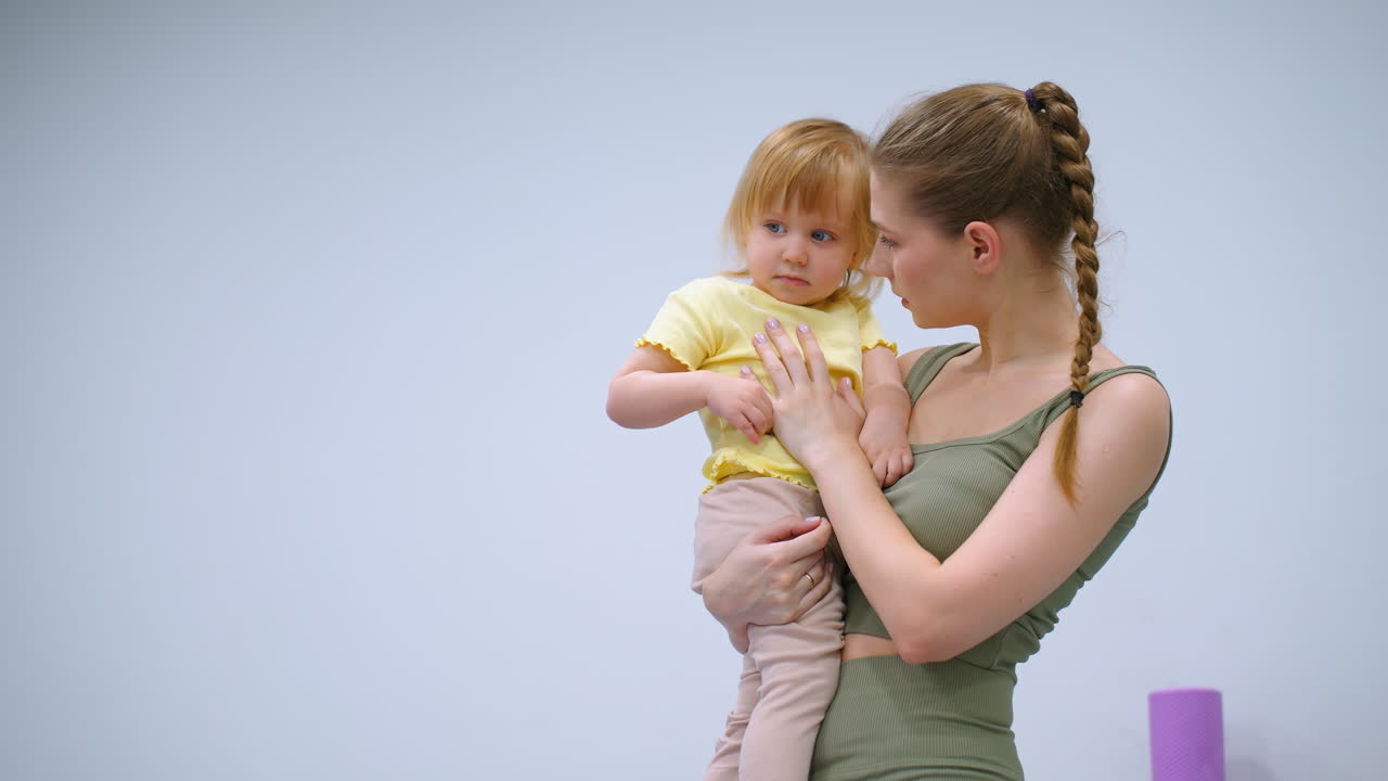 madre en traje verde sosteniendo suavemente a su hija joven en camisa amarilla, ambos de pie en el gimnasio con equipos de yoga púrpura visibles en el fondo