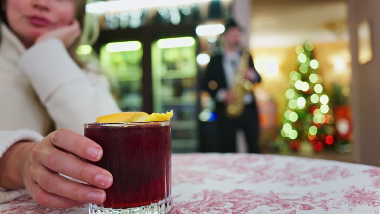 Close up of a woman holding a negroni cocktail on a red and white tablecloth at a restaurant with a Christmas tree on the background