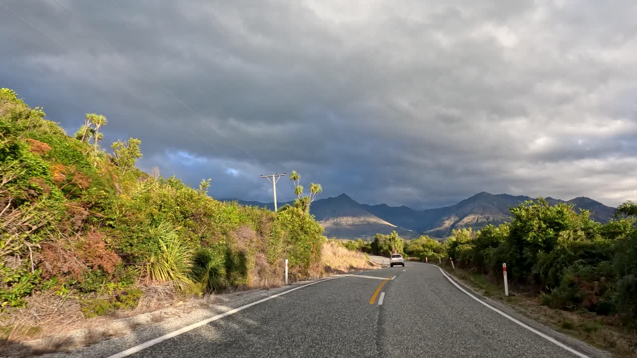 Vehicle travels winding rural road with mountain views, lush greenery, and dramatic cloudy sky lighting